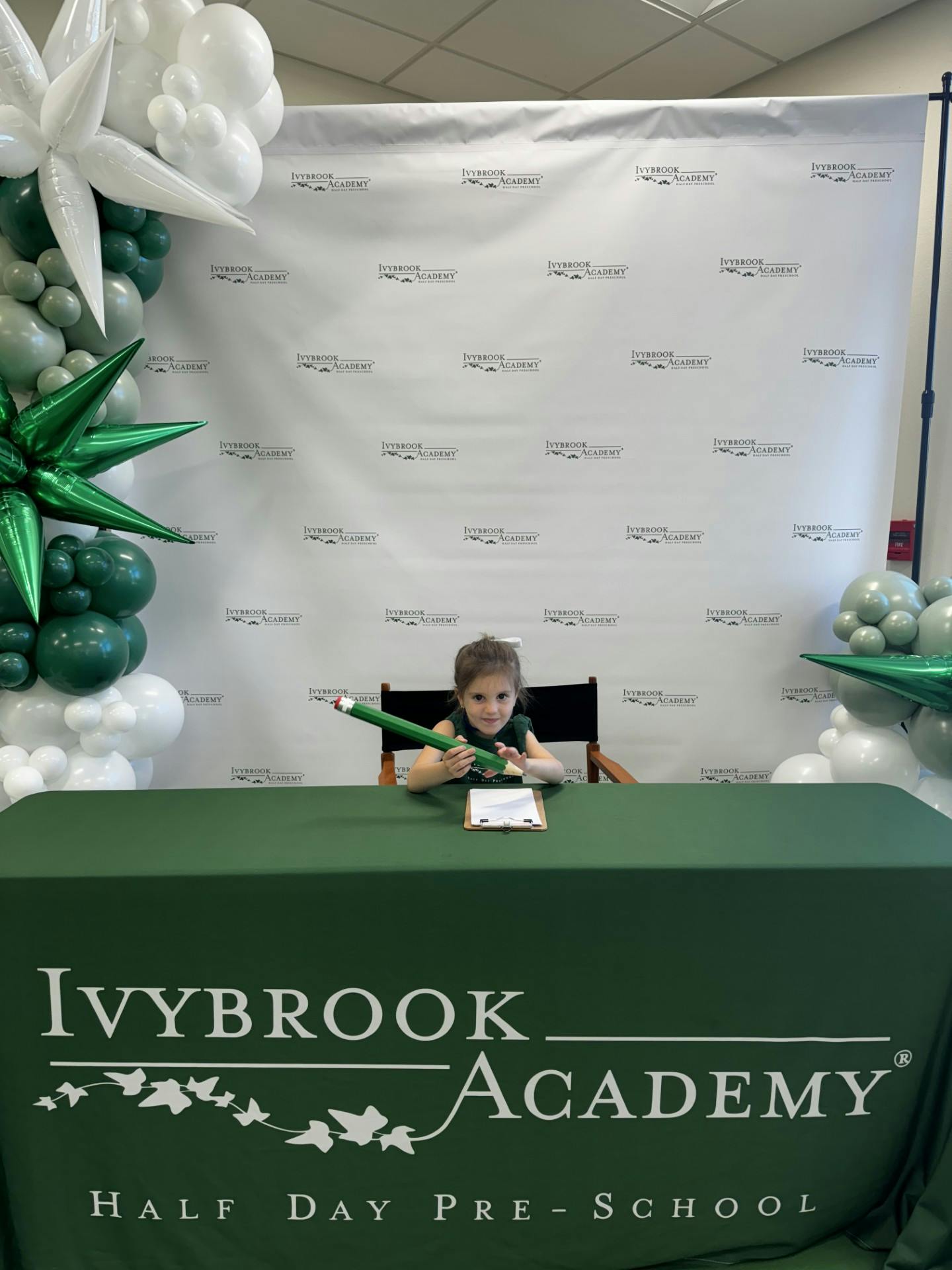 Child sitting at a table with a large green pencil and a clipboard
