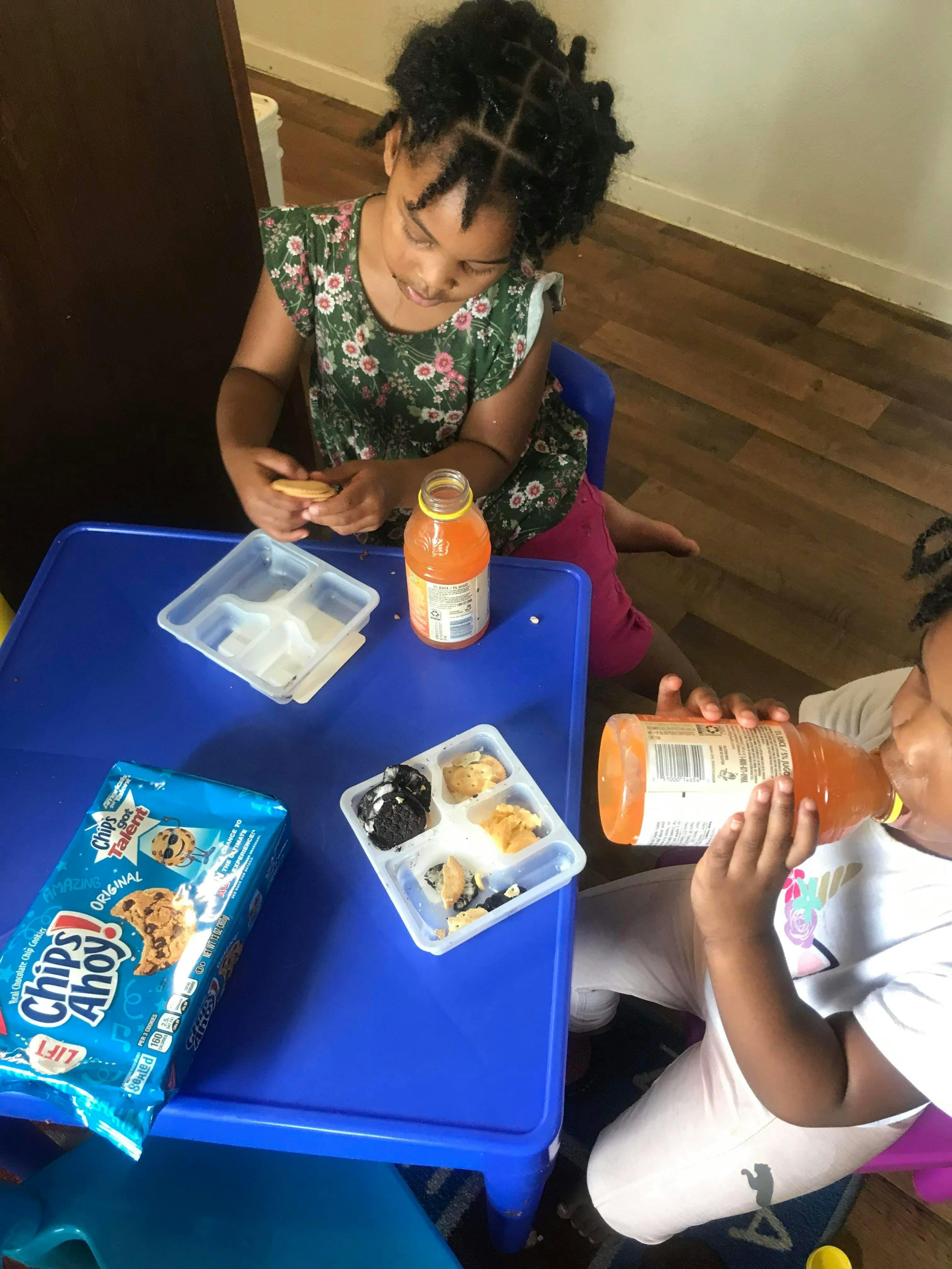 Child eating a cookie at a blue table, another child drinking from a bottle