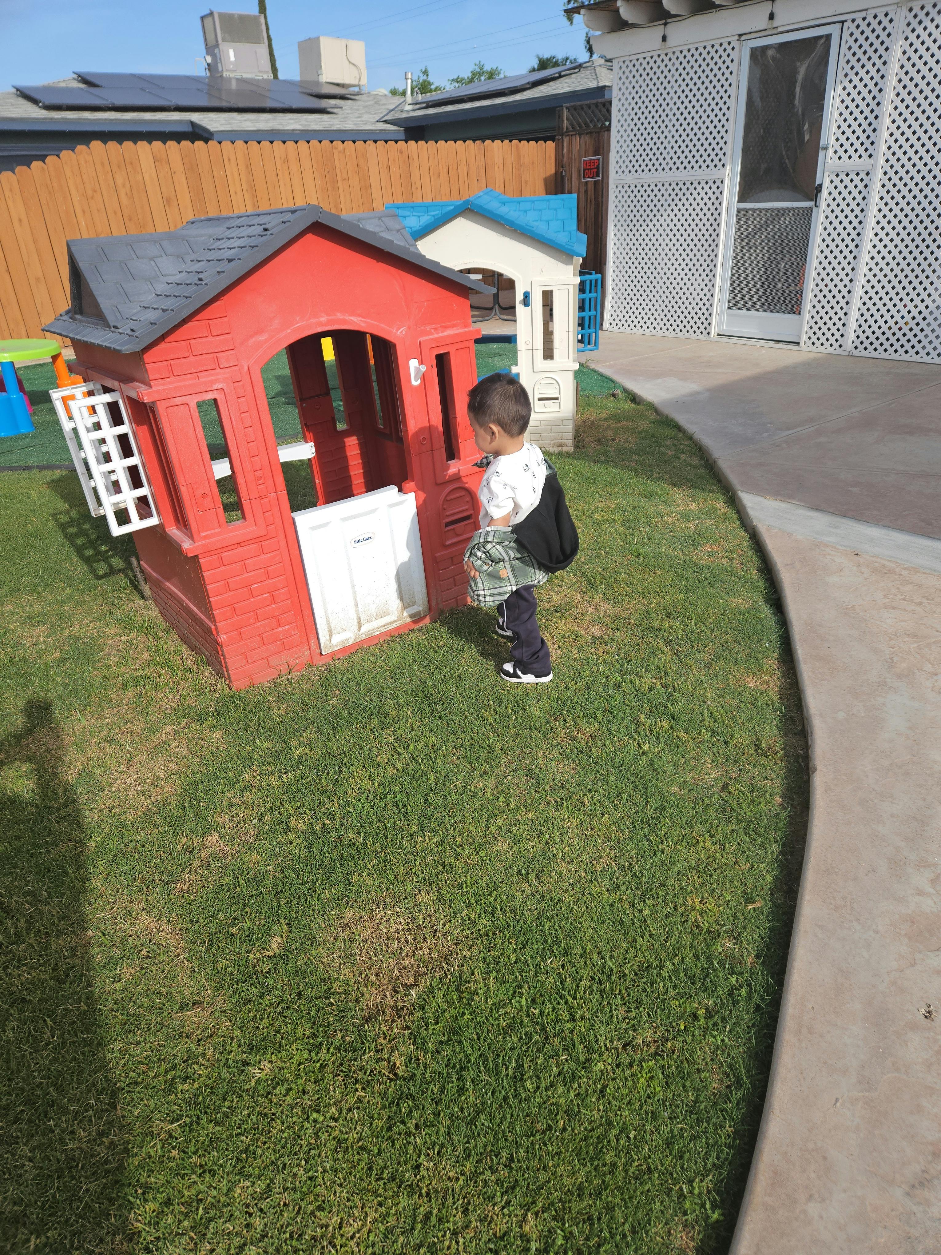 Child bending over a red and white toy playhouse in a grassy area