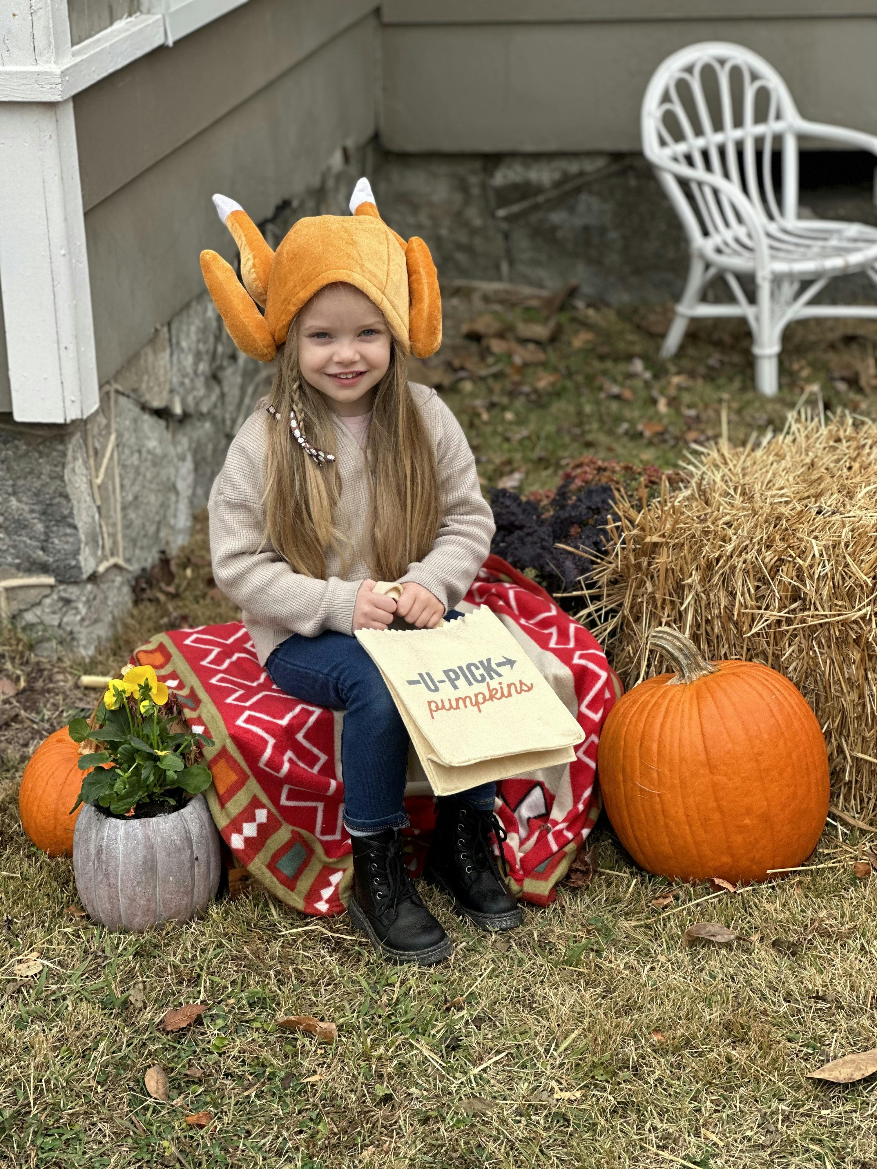 Child in a turkey hat sitting on a blanket next to a pumpkin and hay bale