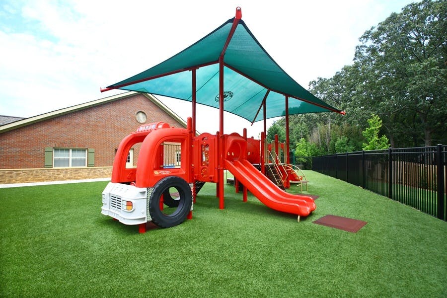 Red playground structure with slides and a green shade canopy on artificial turf