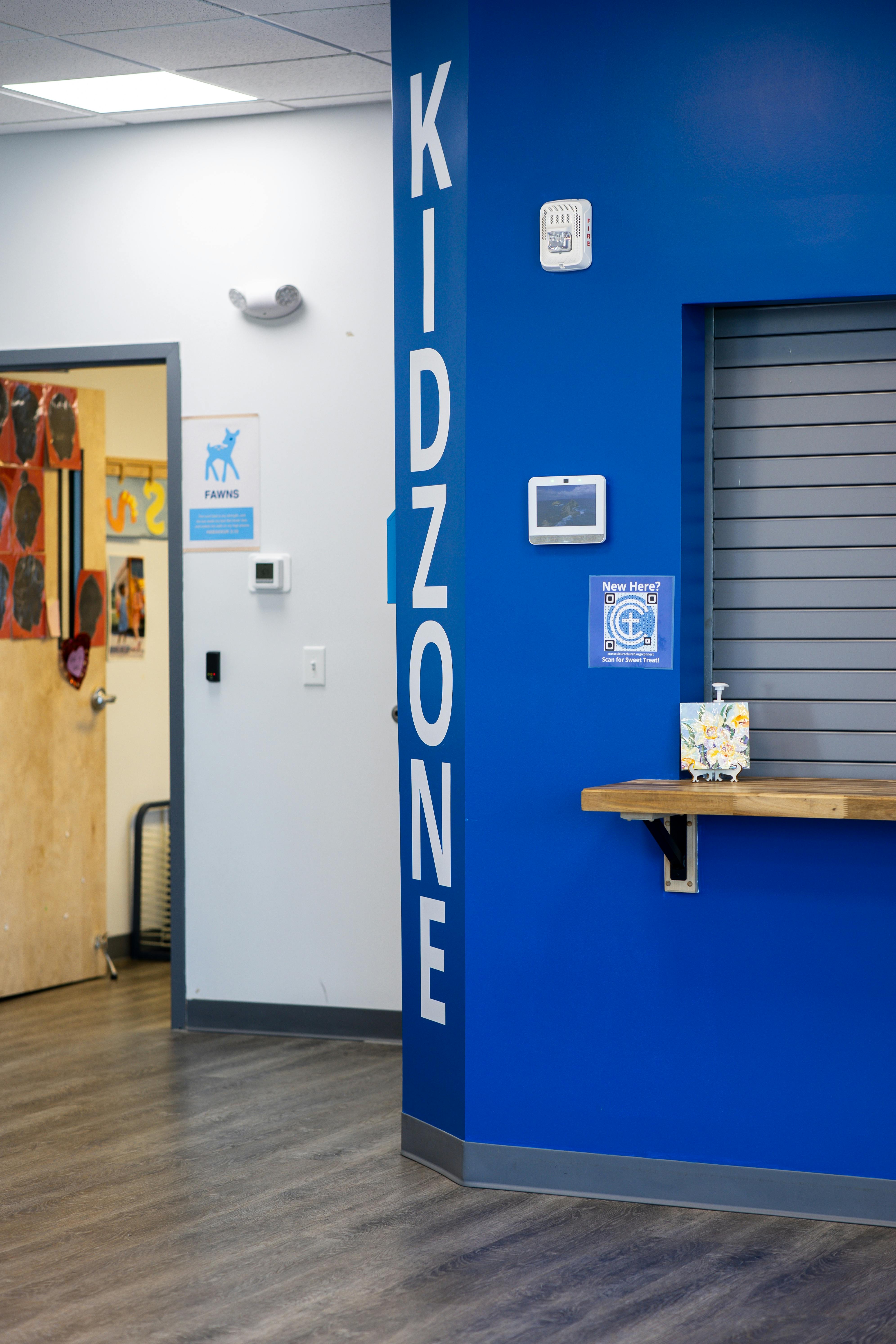 Entrance to a kidzone with a blue wall, desk, and open door to a classroom