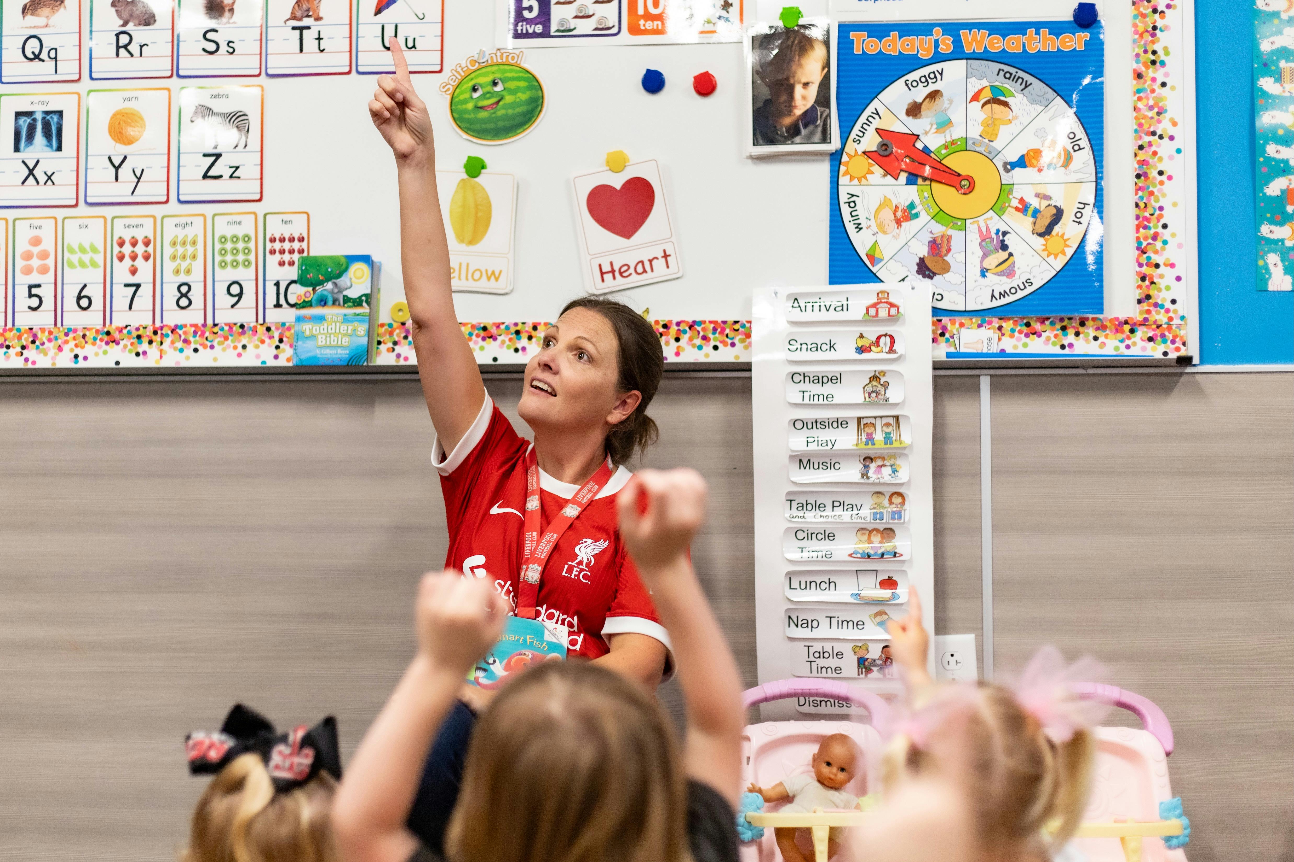 Adult in red shirt with arm raised, children with arms raised in classroom