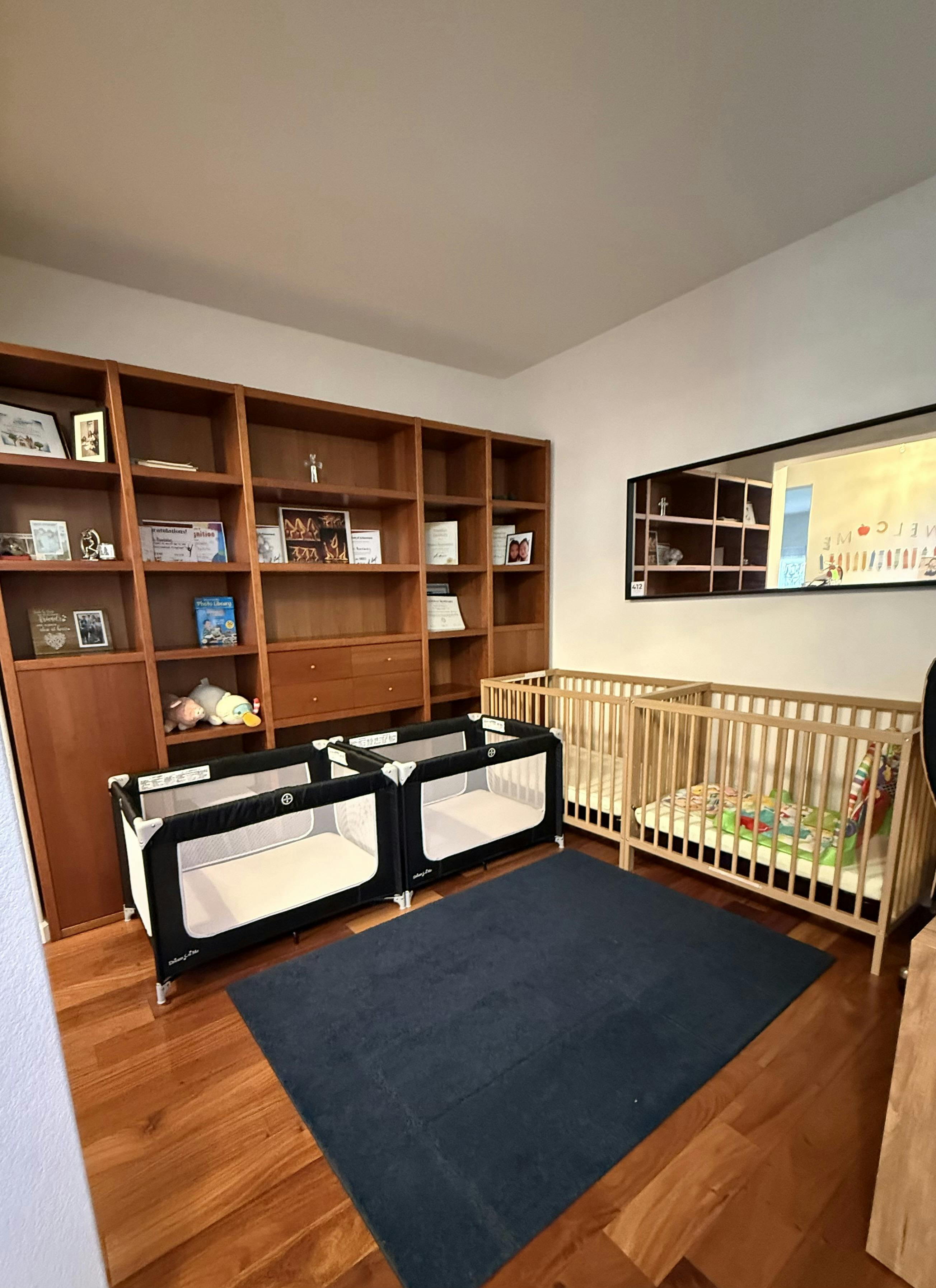 Two black portable cribs and two wooden cribs on a rug in a room with shelves