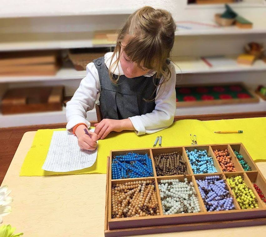 Child writing on paper at a table with a box of colorful beads