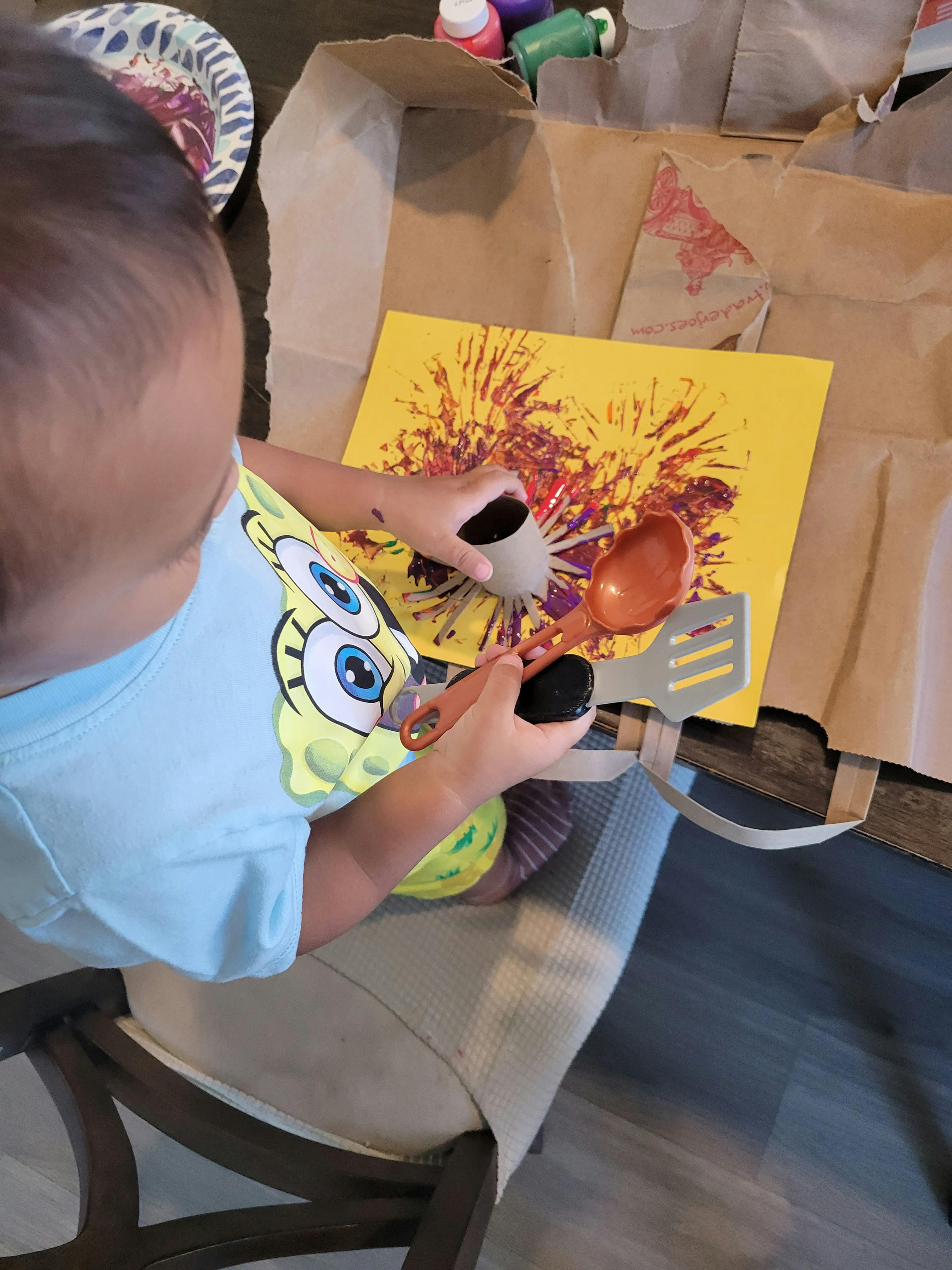 Child painting on yellow paper using a cardboard roll and toy kitchen utensils