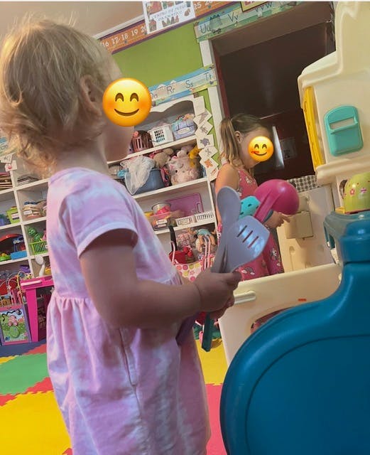 Child holding toy utensils, another child playing with a toy kitchen