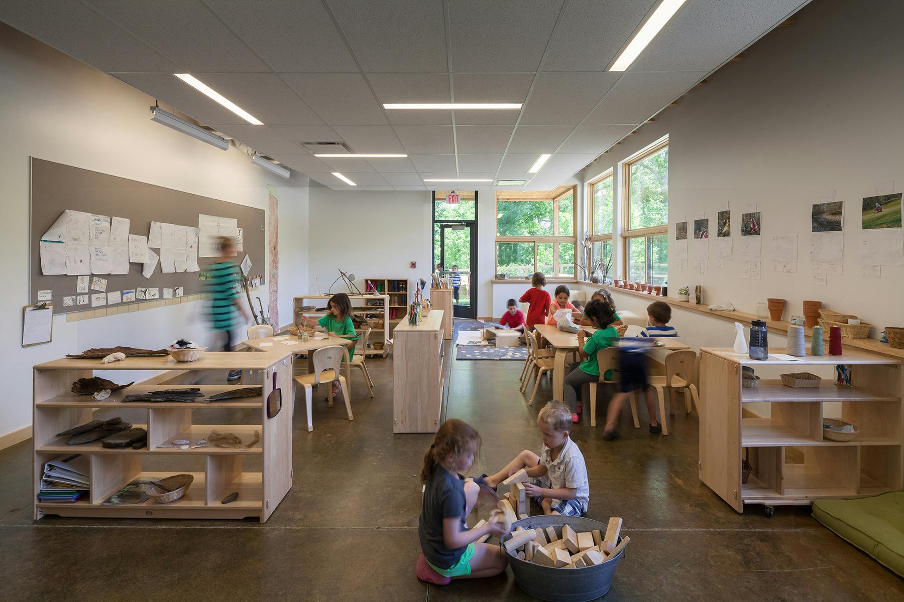 Children in a classroom with tables, shelves, and large windows