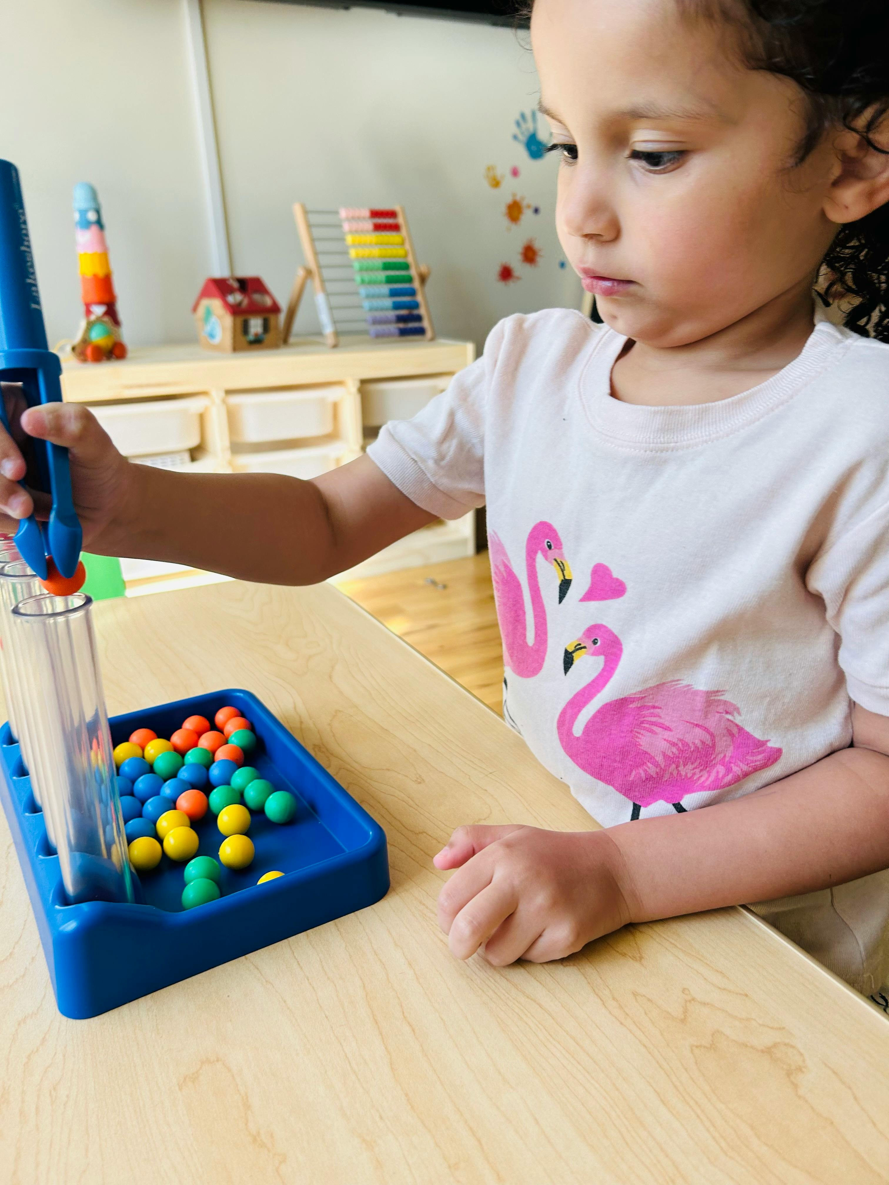 Child using a tool to pick up a colorful ball from a tray of balls