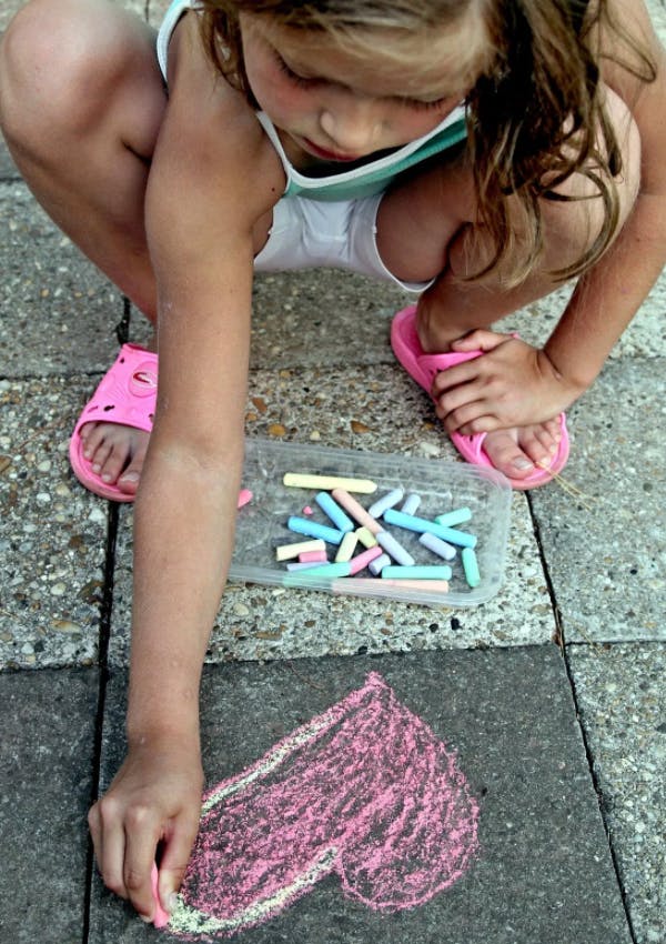 Child drawing with pink chalk on pavement next to a container of chalk