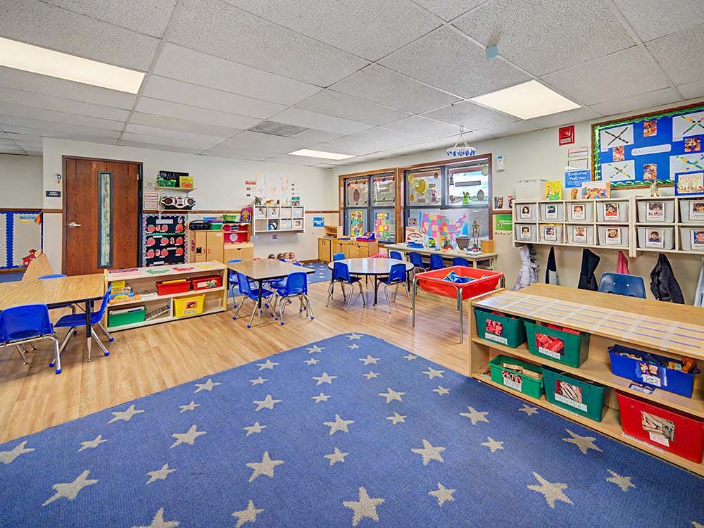 Classroom with small tables and chairs, shelves with toys, and a star-patterned rug