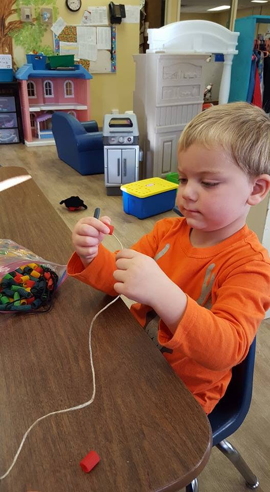 Child stringing beads at a table in a classroom