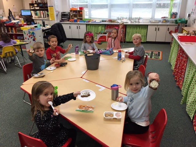 Children sitting at a table eating snacks in a classroom, some holding up food