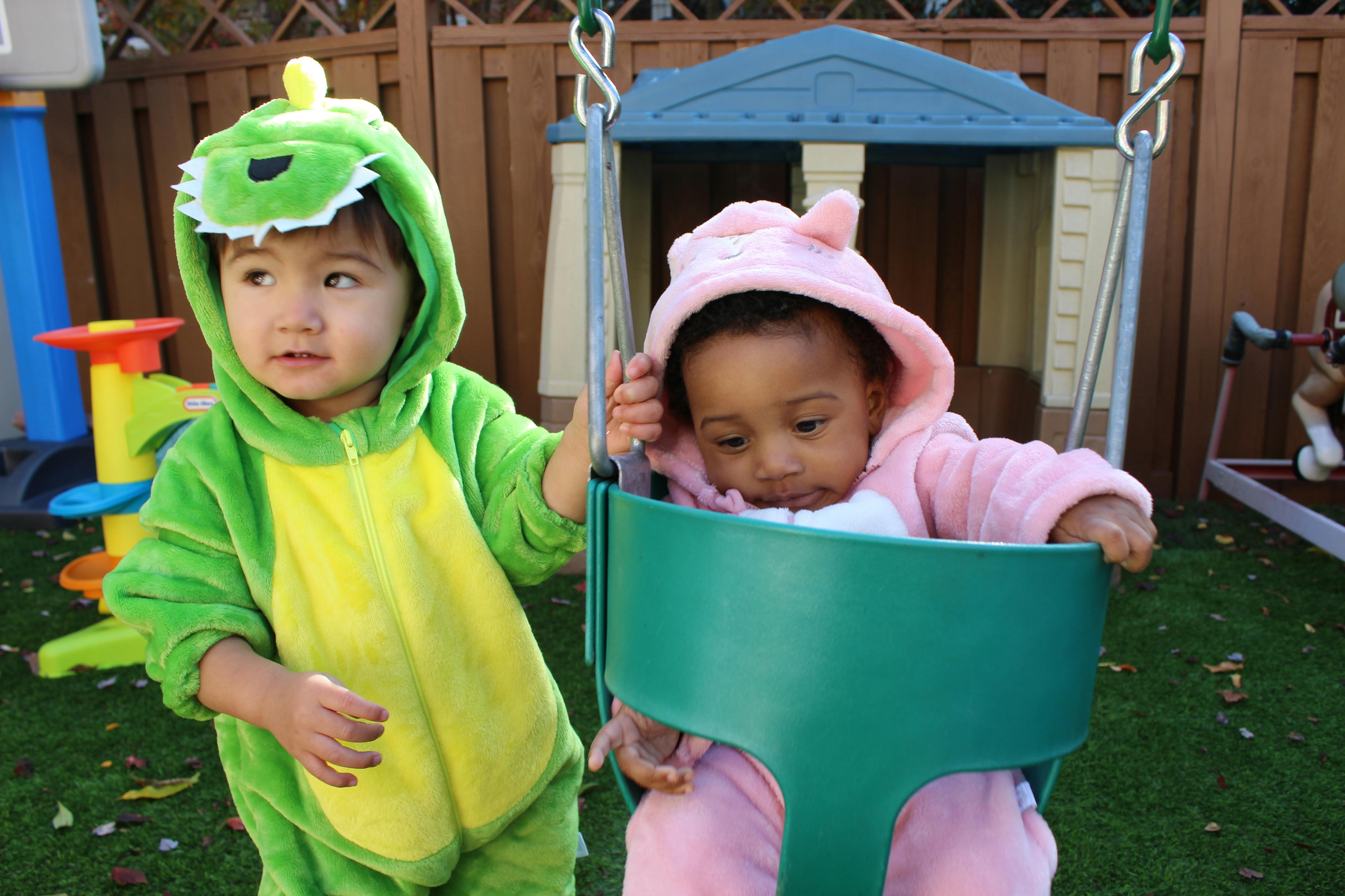 Child in dinosaur costume next to child in swing