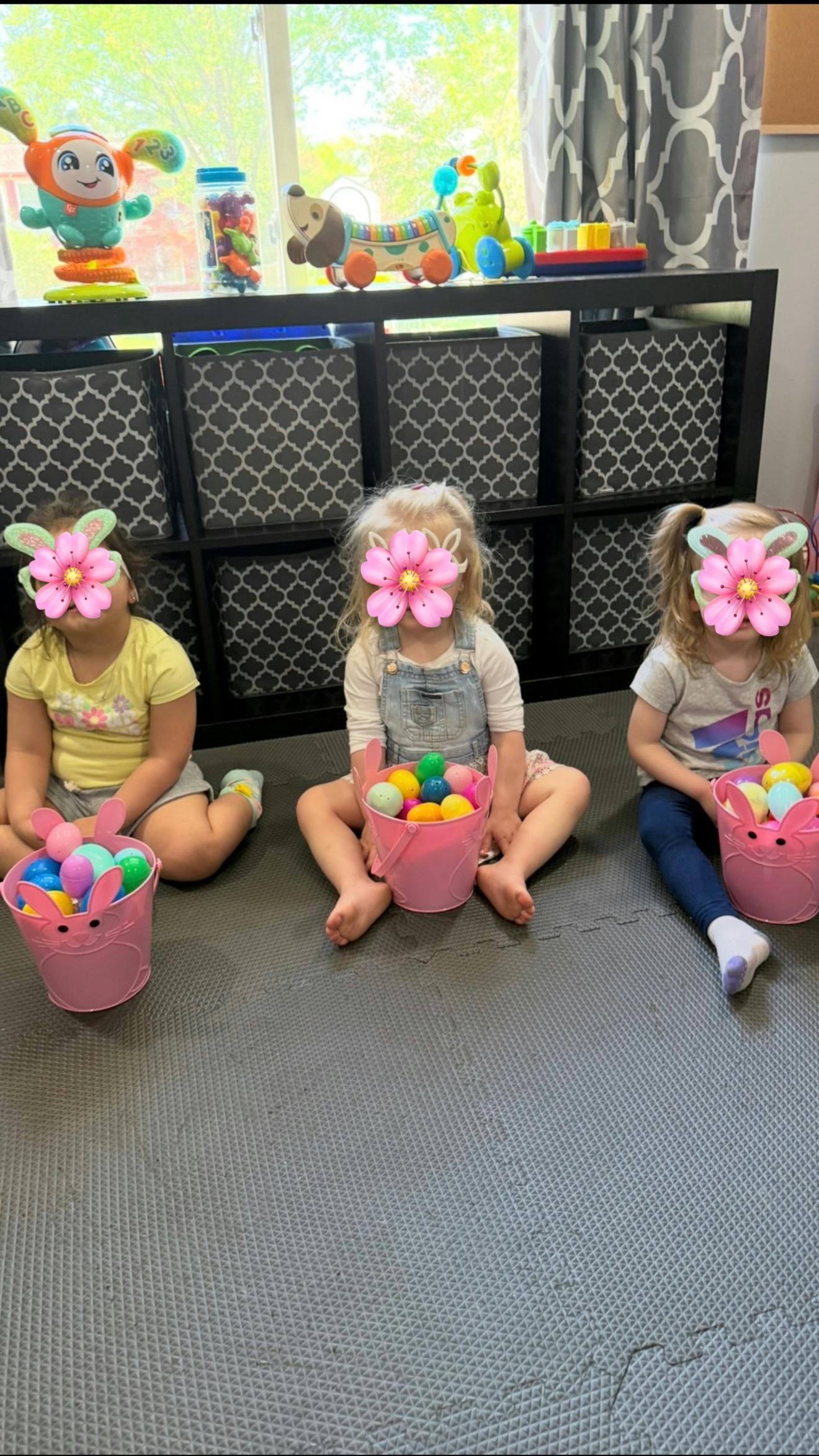Three children sitting on a mat with pink buckets filled with colorful plastic eggs