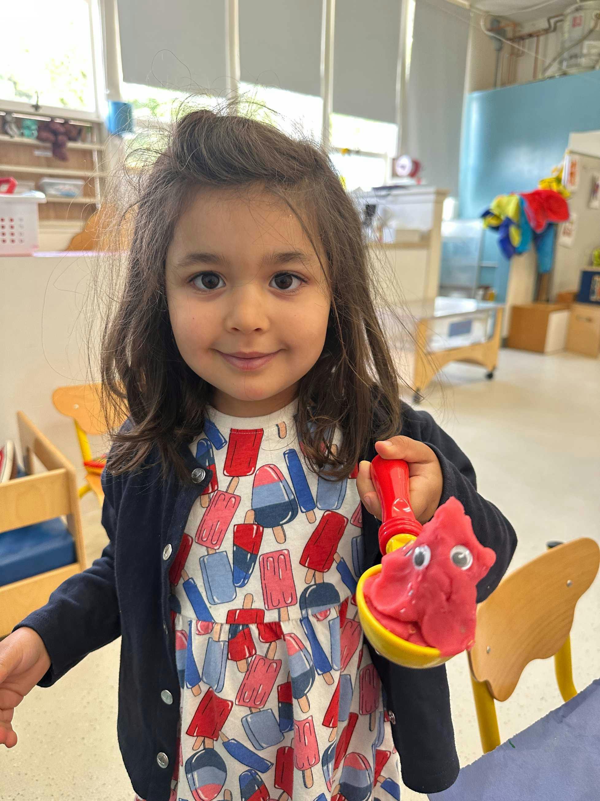Child holding a red play-doh creation with googly eyes in a yellow scoop