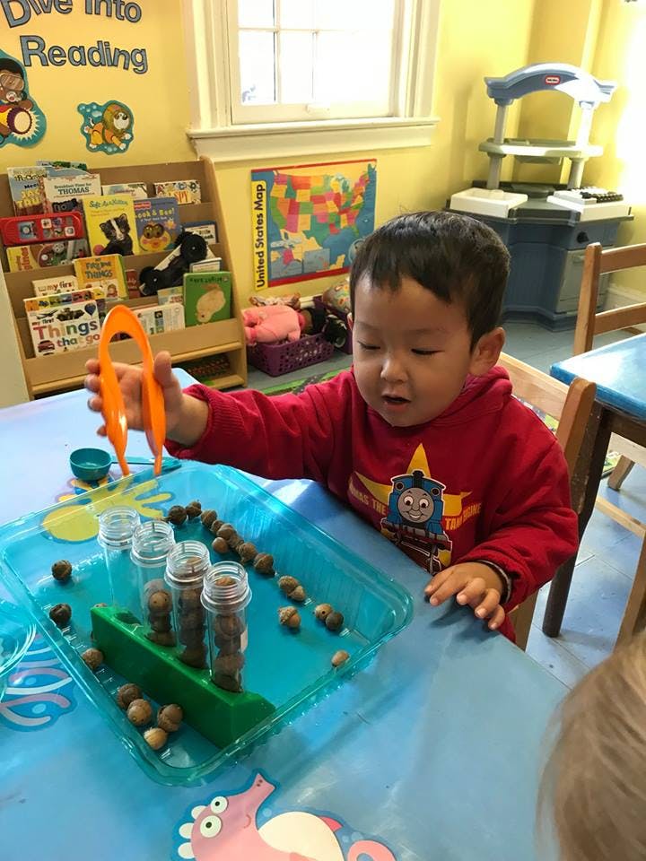 Child using orange tongs to pick up small objects in a classroom
