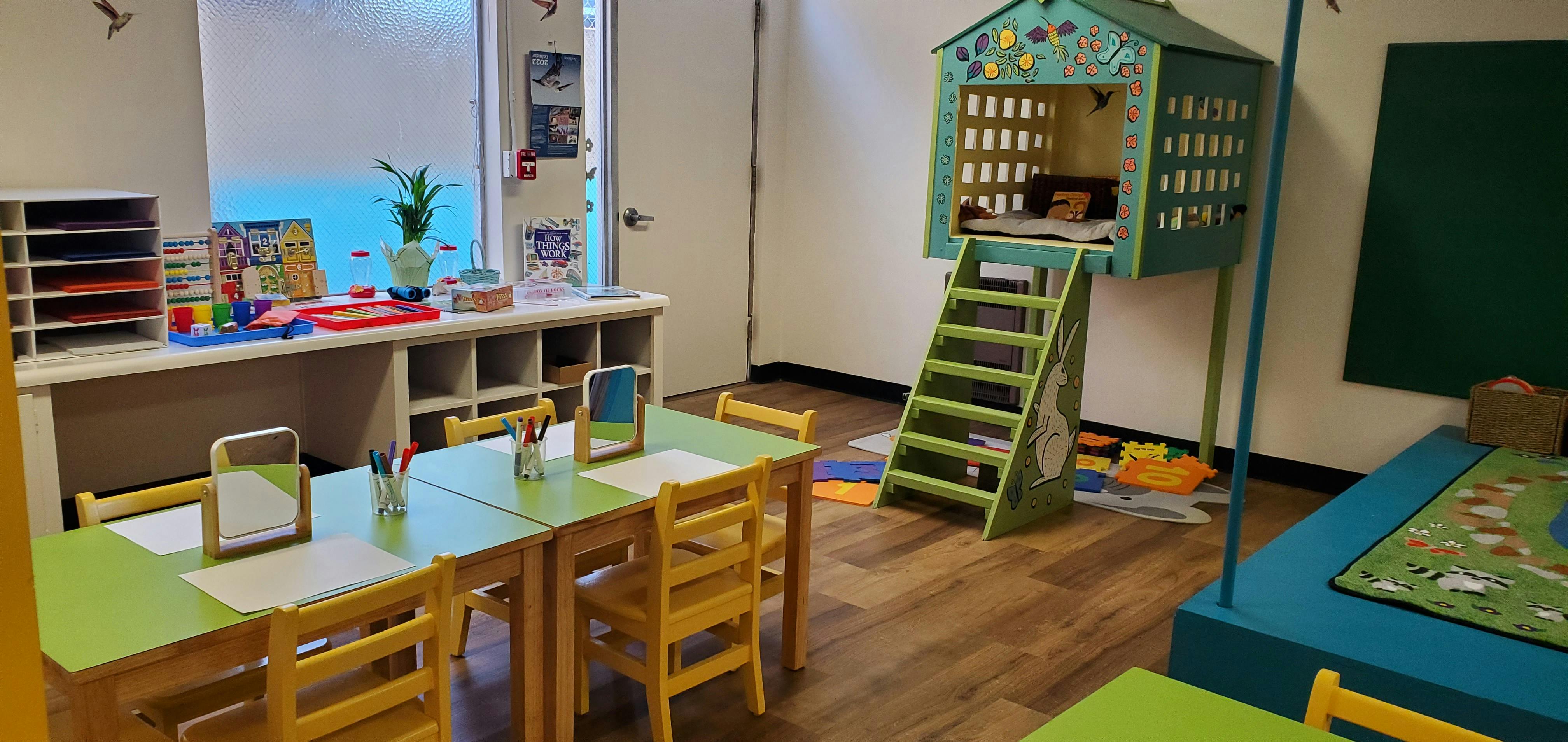 Classroom with small tables and chairs, and a playhouse with a ladder