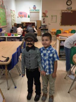 Two children standing in a classroom, one in costume with a fake mustache