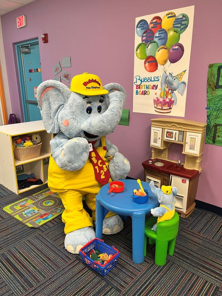 Elephant mascot in yellow overalls and hat standing next to a small blue table