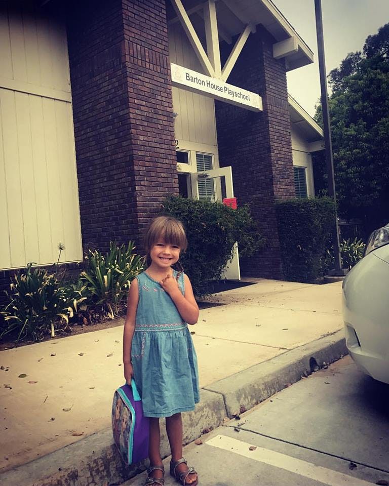 Child in a dress holding a lunchbox in front of a building with a sign