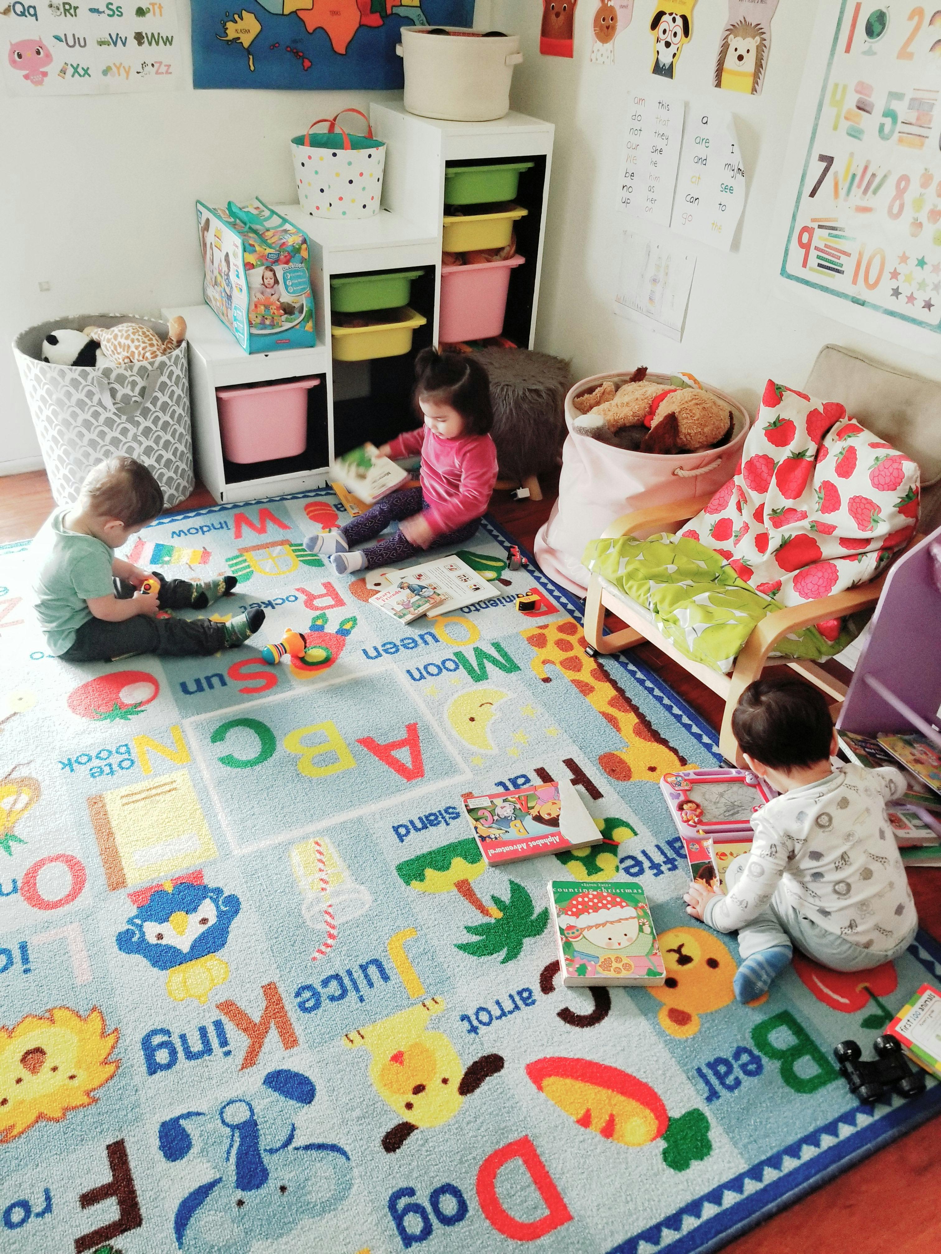 Three children playing with books and toys on an alphabet rug in a playroom