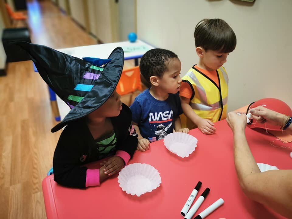 Three children at a table, one in a witch hat, one in a NASA shirt, one in a safety vest