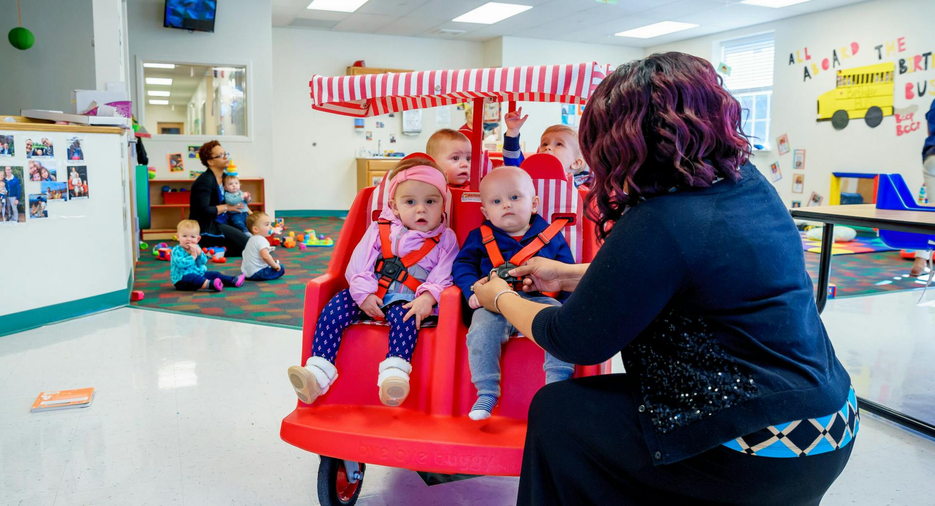 Person helping children in a multi-child stroller in a classroom