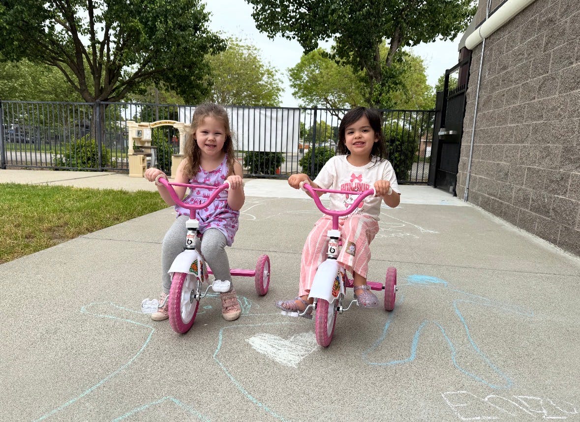 Two children riding pink tricycles on a paved surface with chalk drawings