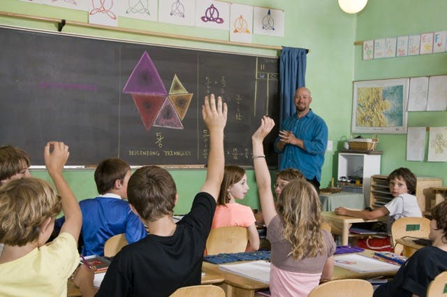 Children raising hands in a classroom with an adult standing by a chalkboard