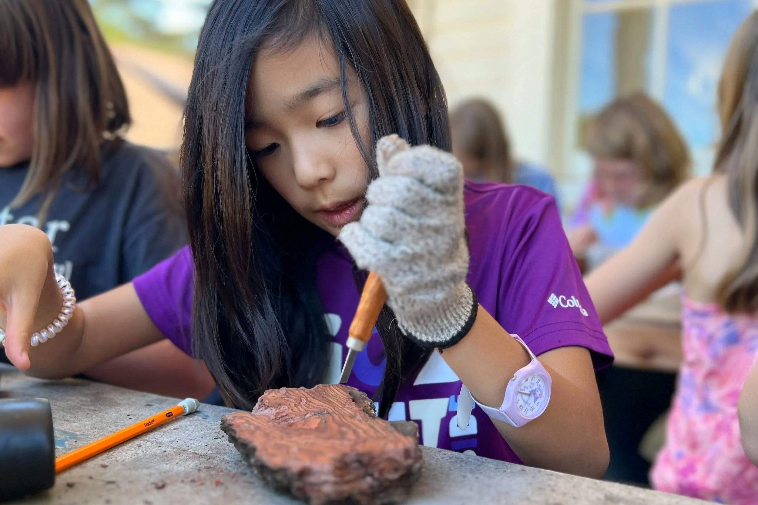 Child carving a piece of wood with a tool and knife, wearing a glove