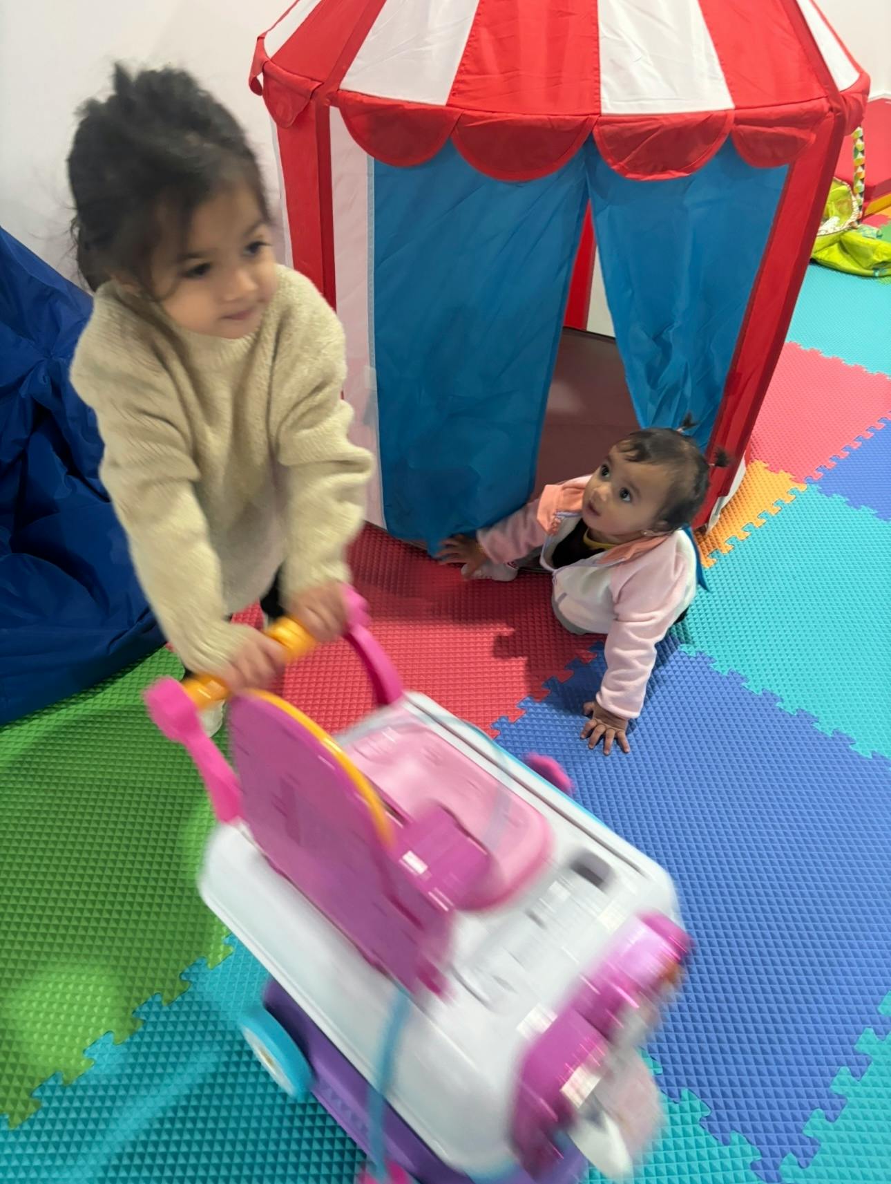 Child pushing a toy stroller and a baby crawling out of a play tent