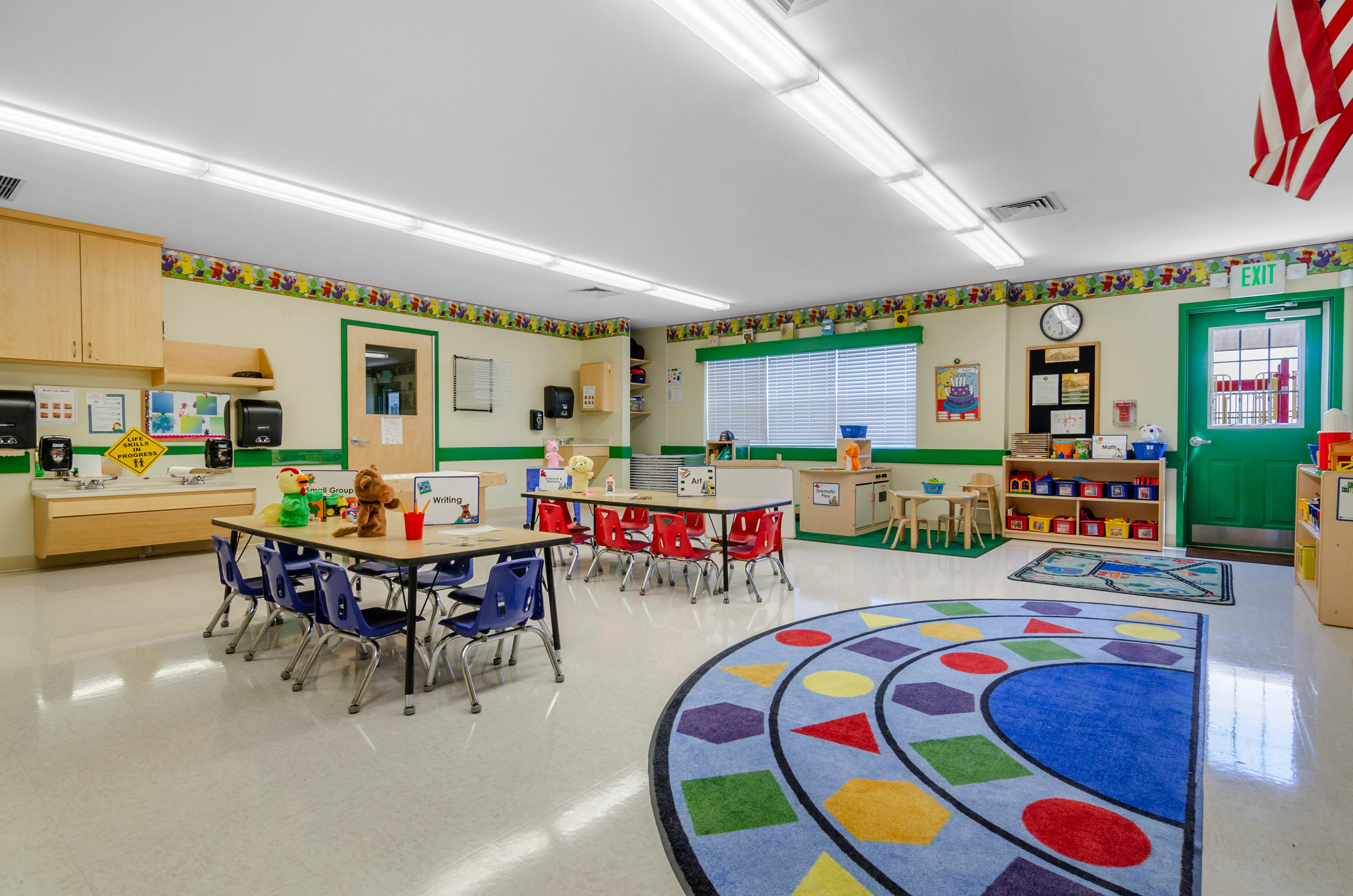 Classroom with child-sized tables and chairs, wall-mounted sinks, and a shape rug