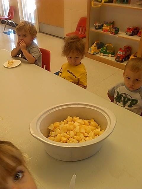 Three children sitting at a table with a bowl of diced fruit