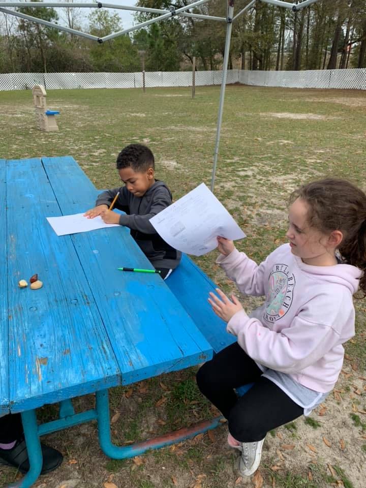 Two children at a blue picnic table, one writing and one holding paper