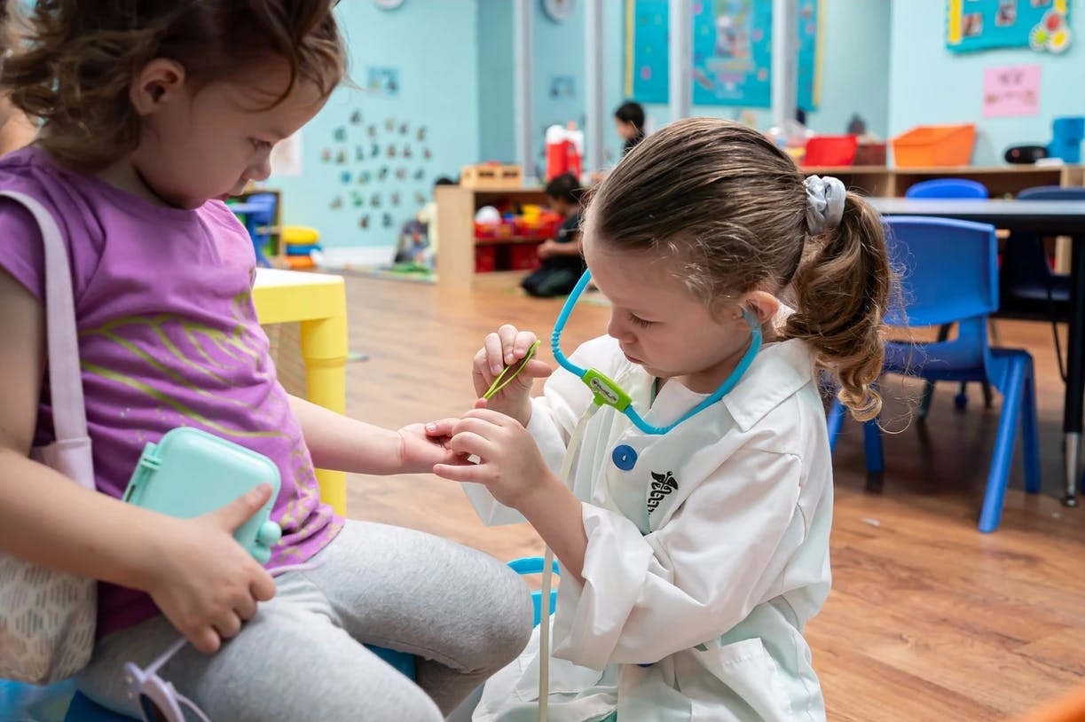 Child in white coat playing doctor with another child in a classroom