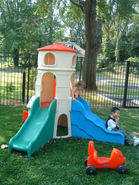 Child on a blue slide next to a play structure, another child sitting on grass