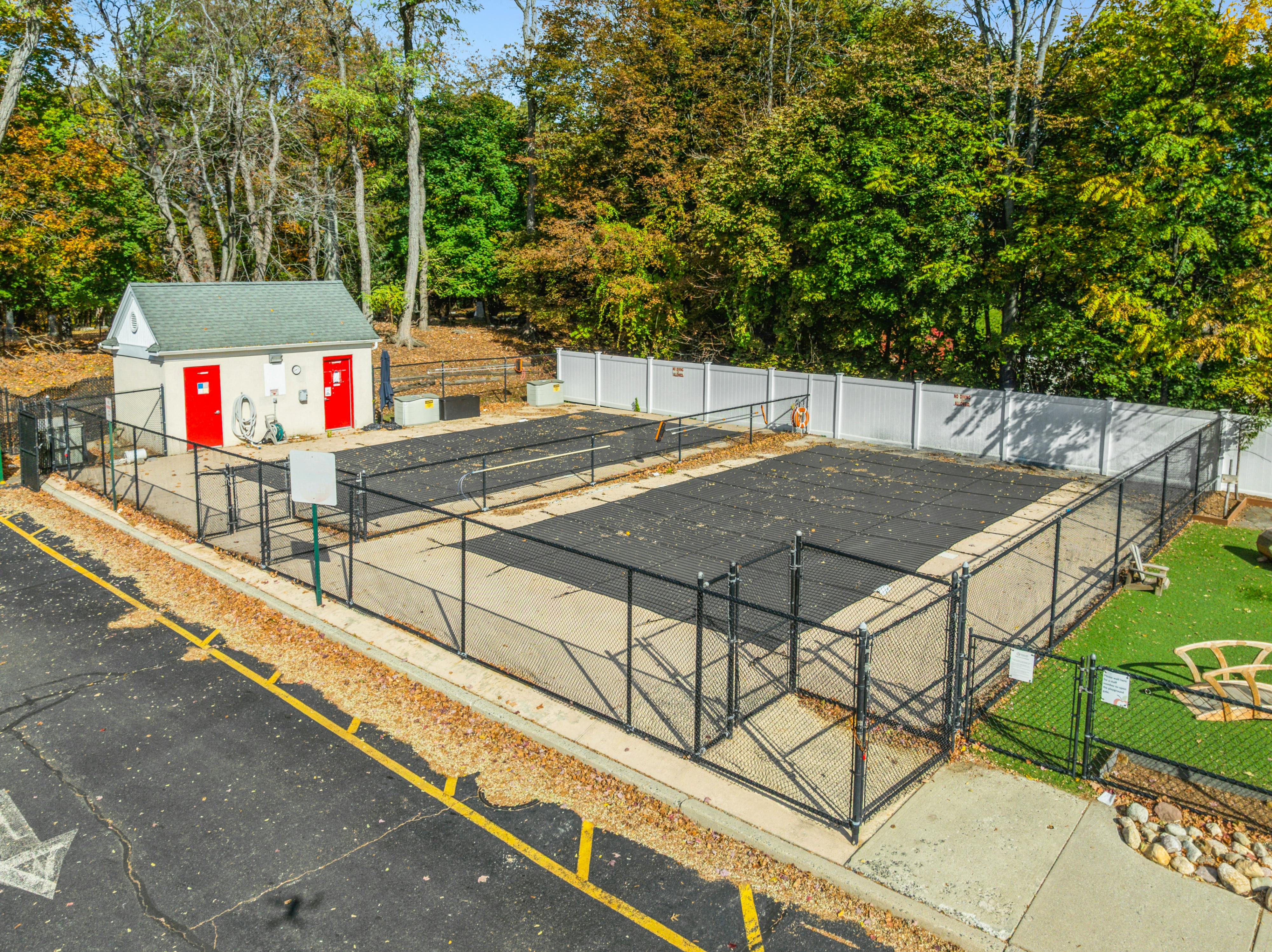 Fenced-in swimming pool area with a small building and parking lot in autumn