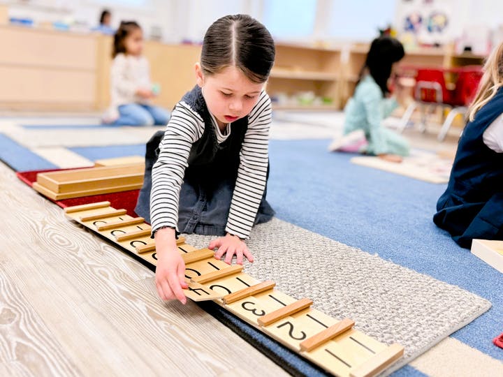 Child on floor playing with wooden number puzzle inlays, other children in background