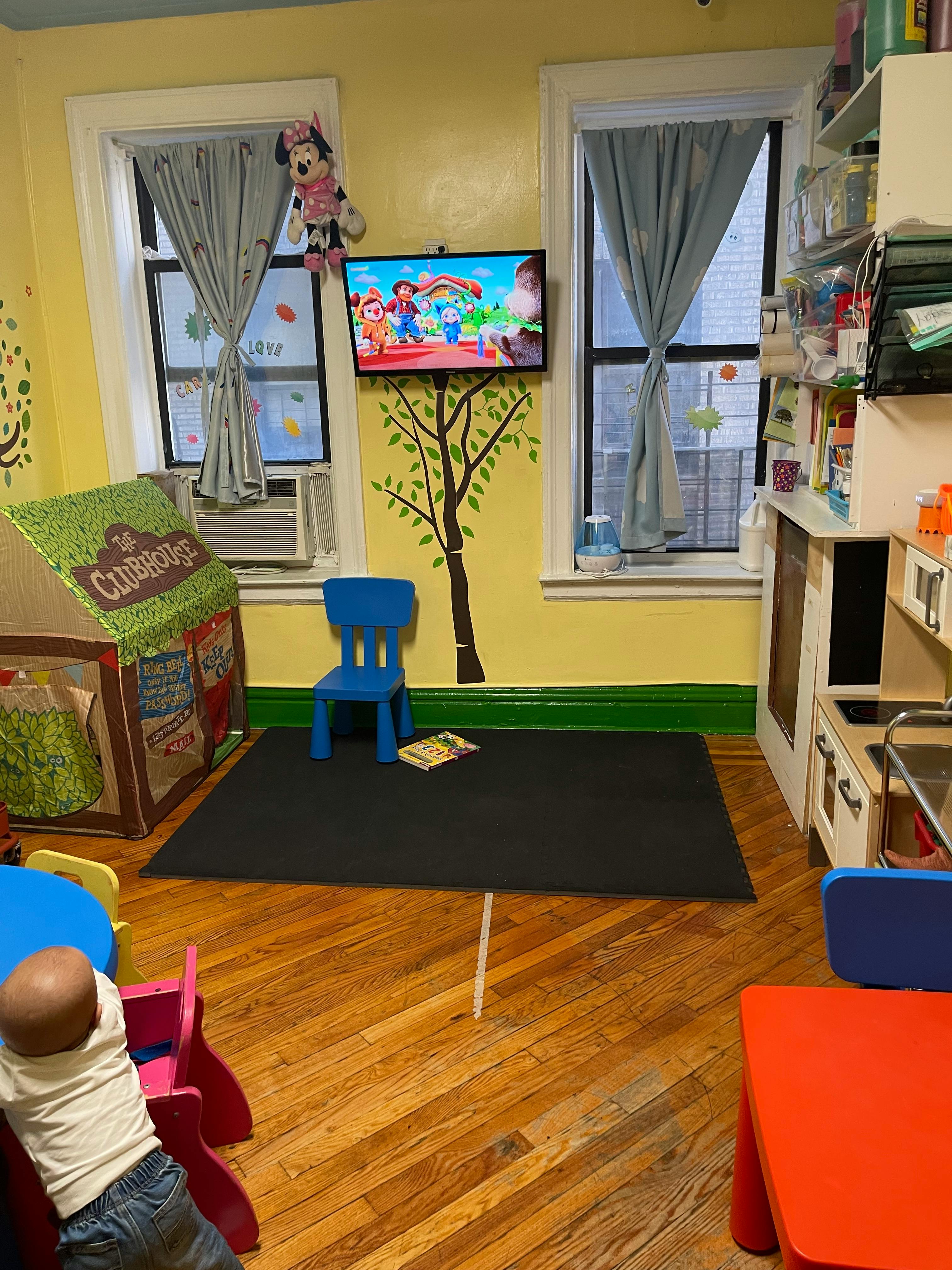 Child standing near a blue table in a playroom with a TV playing cartoons