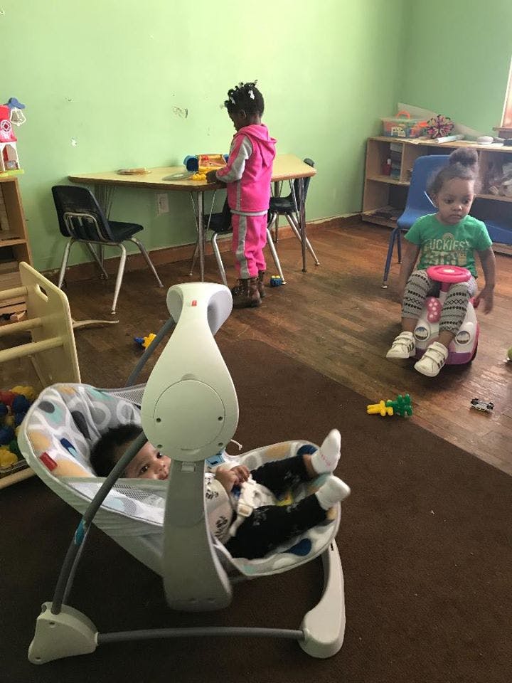 Three children in a room, one in a baby swing, one at a table, one on a ride-on toy