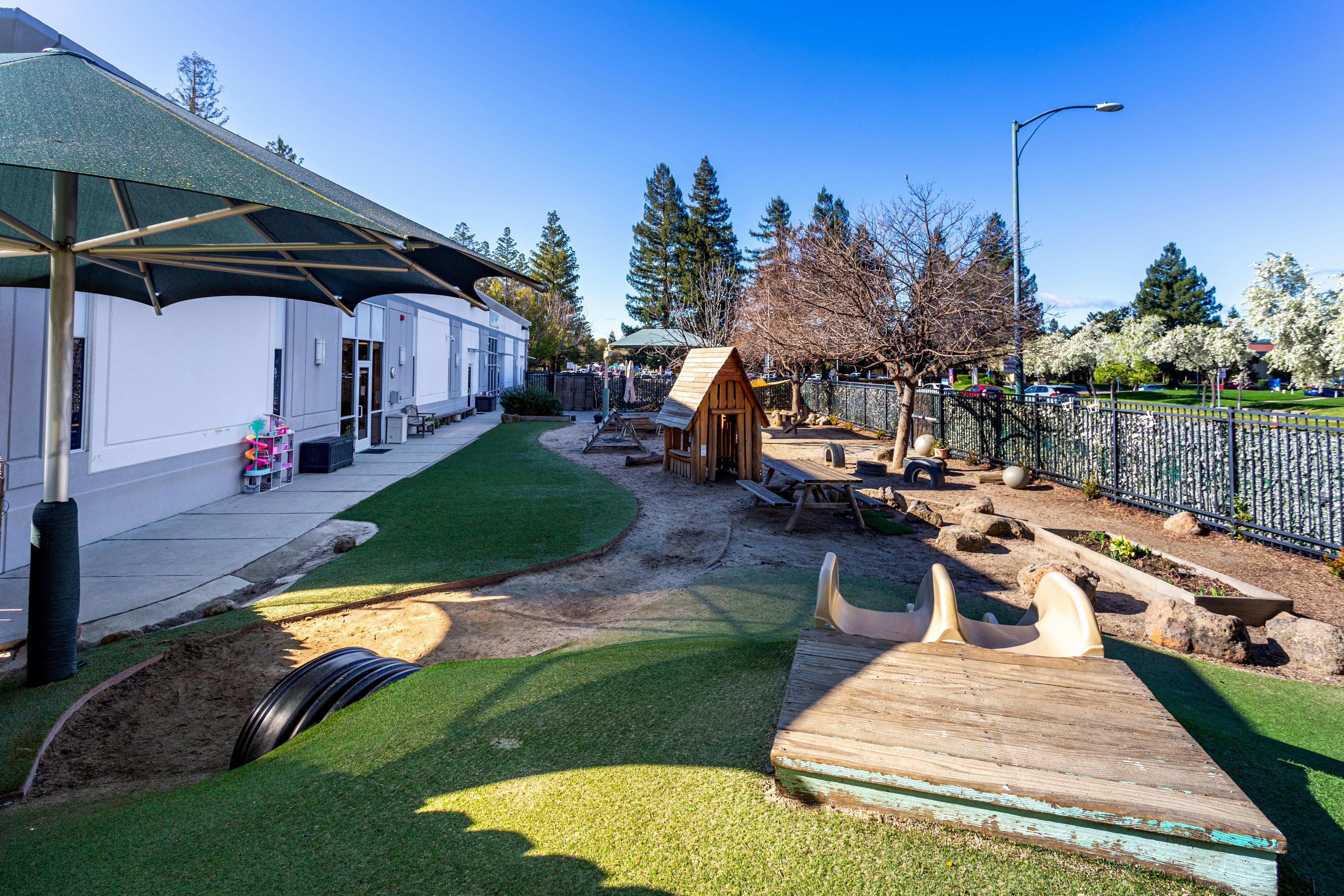 Outdoor playground with artificial turf, wooden play structures, and a building