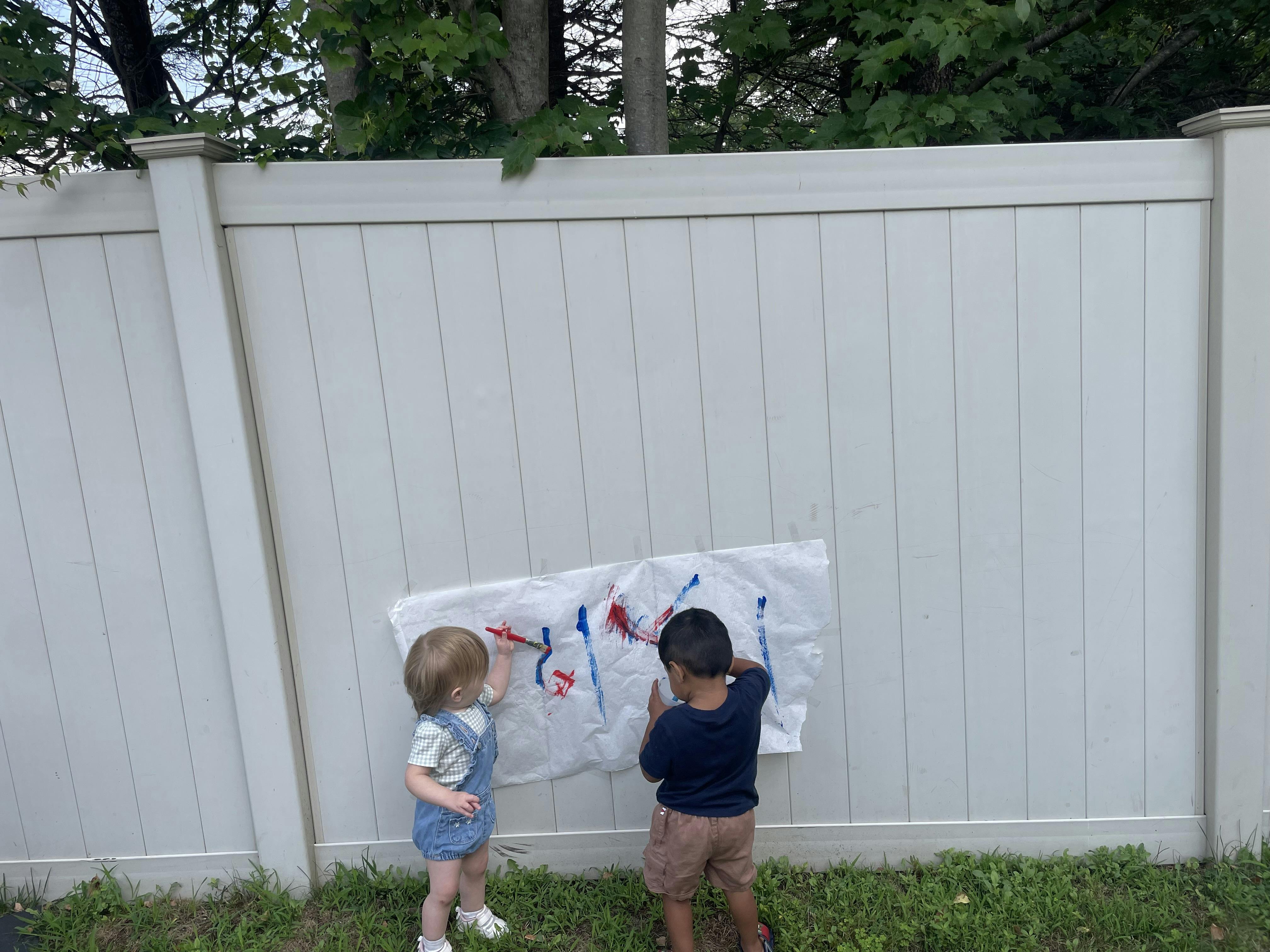 Two children painting on paper attached to a white fence