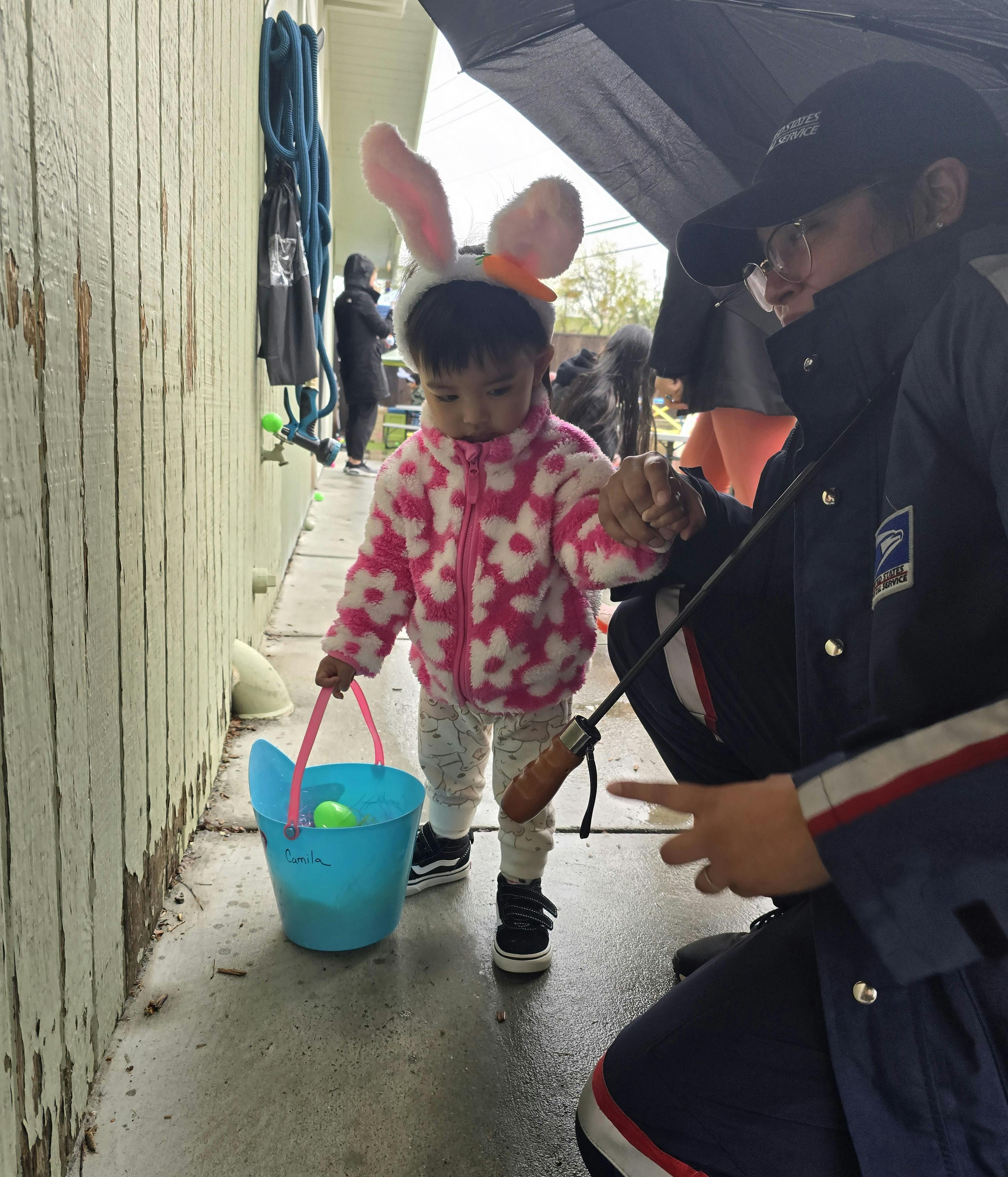 Child in bunny ears holding a bucket with a person under an umbrella