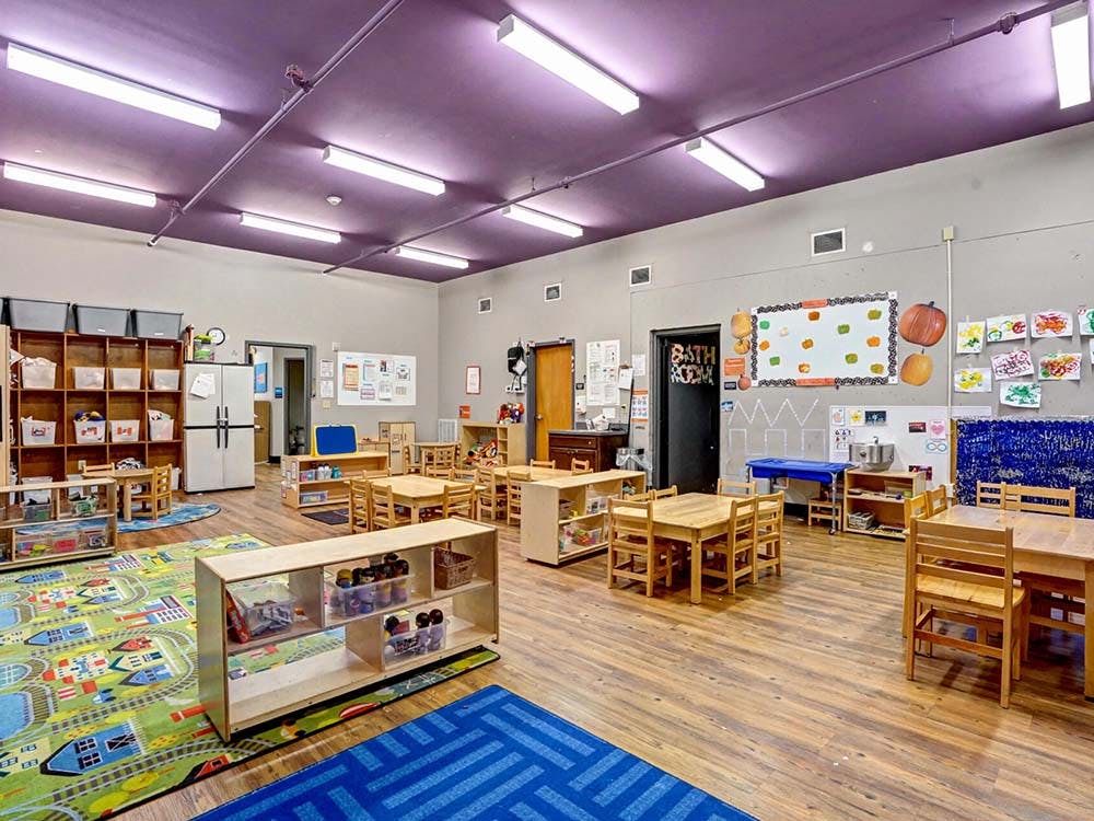 An empty classroom with small wooden tables and chairs, play mats, and cubbies