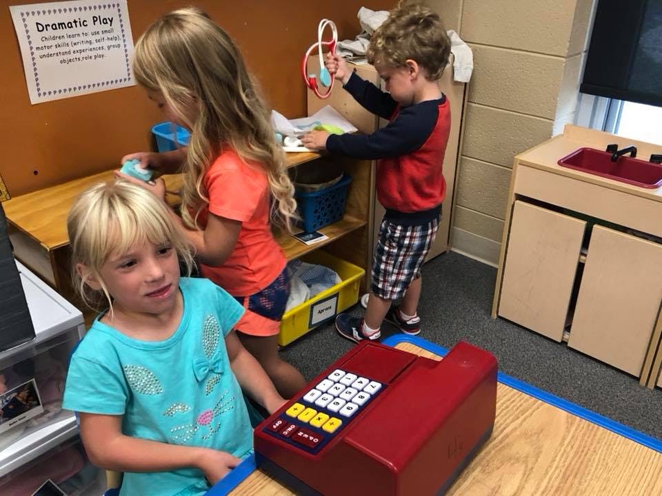 Three children playing in a classroom with a toy cash register