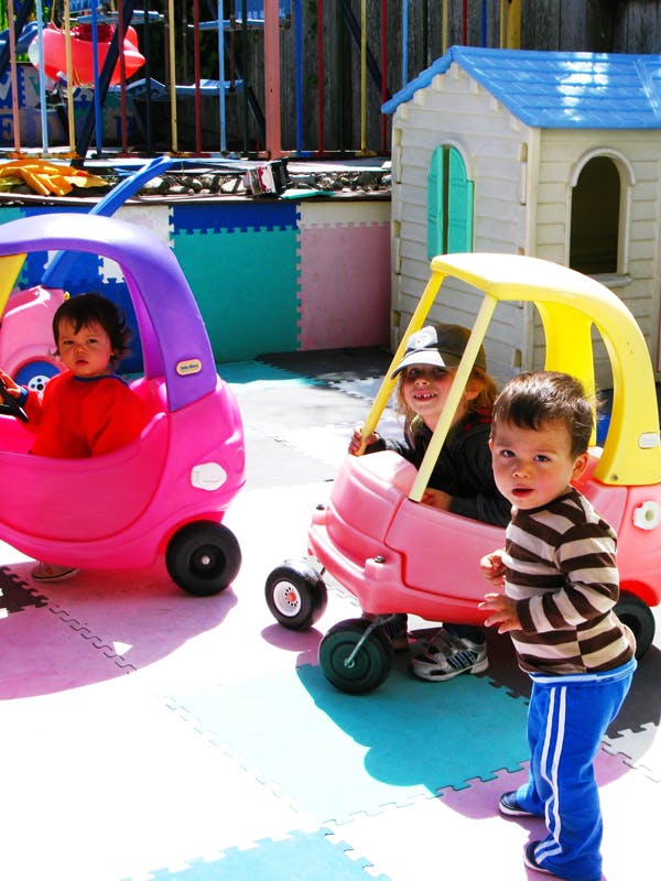 Three children playing outdoors with toy cars and a playhouse