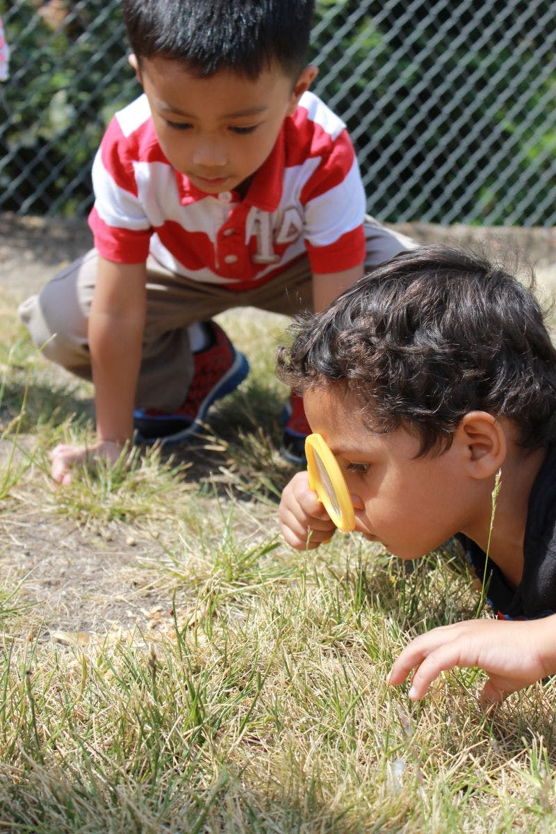 Child looking through a magnifying glass at the ground, another child crouching