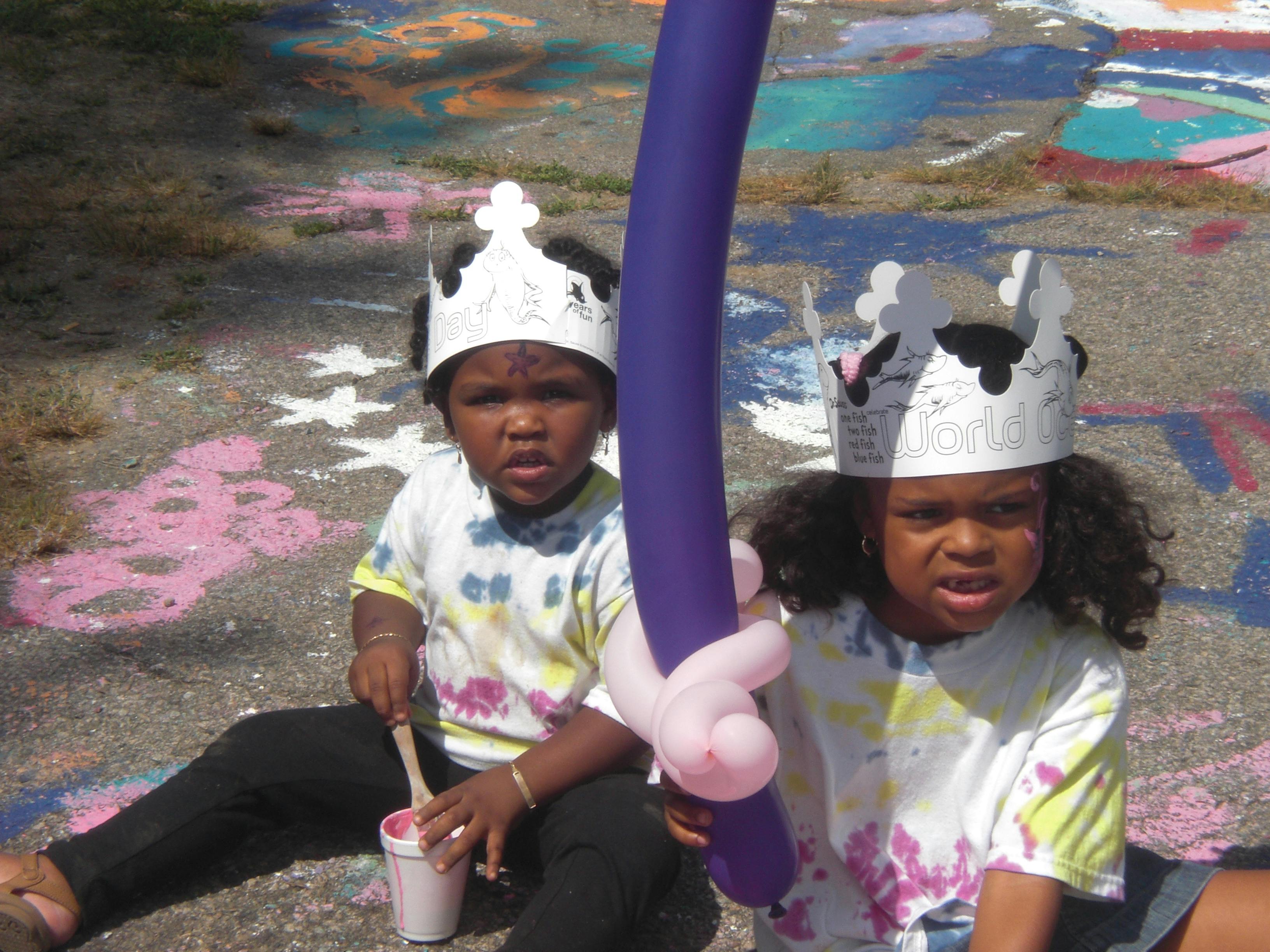 Two children wearing crowns sitting on pavement with a balloon sword