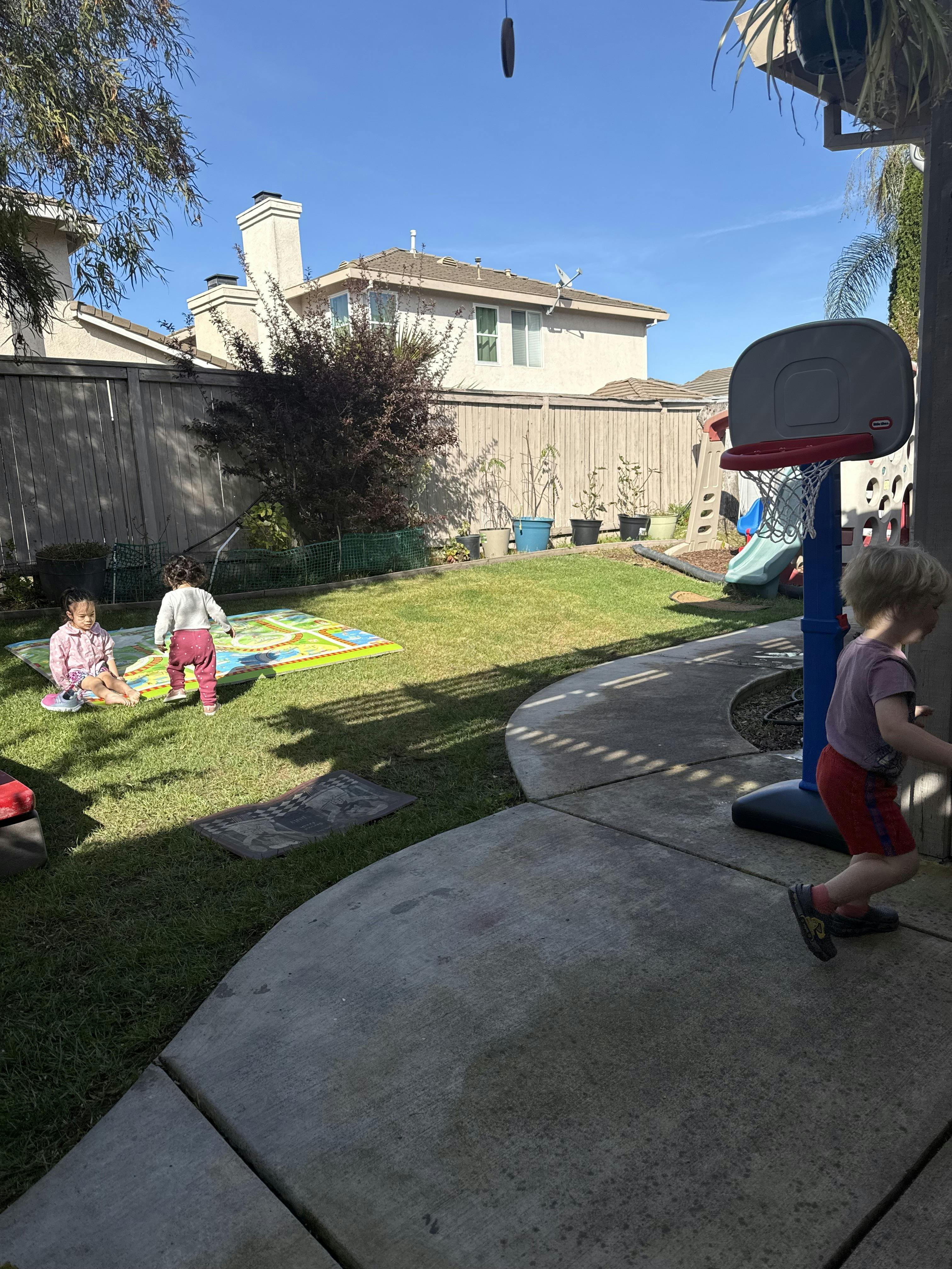 Three children playing in a backyard, one child walks past a toy basketball hoop